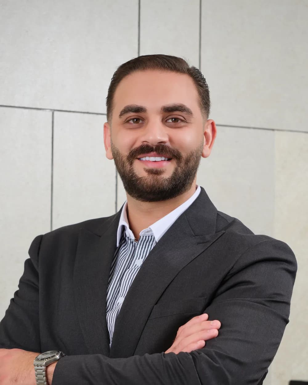 Jamal AlKayid in a charcoal suit, arms crossed, against a stone-tiled wall