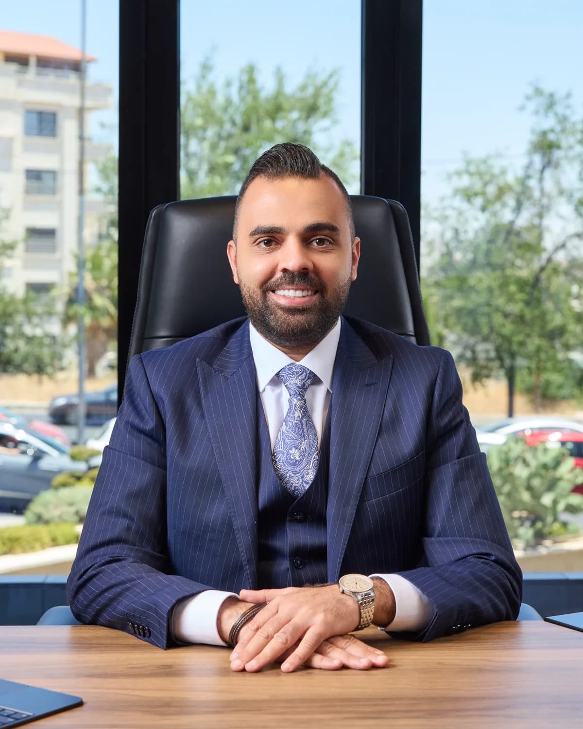 Rami Musallam in a blue suit, seated at a desk in front of floor-to-ceiling office windows